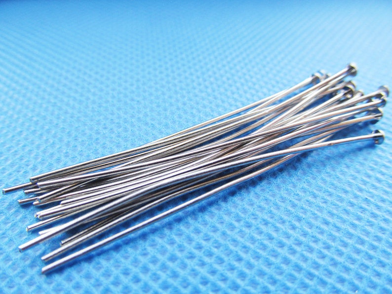 May include: A bundle of silver-colored, straight, thin metal pins with rounded ends. The pins are arranged in a loose pile on a blue textured surface.