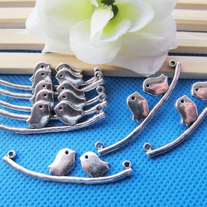 May include: Silver tone metal bird charms on a branch design. The charms are in the shape of birds with a simple design. The branches are silver tone metal and have a loop at the end for attaching to a necklace or bracelet.