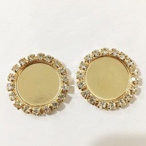 May include: Two gold-toned round metal settings with clear rhinestone borders. The settings are blank and can be used for crafting.