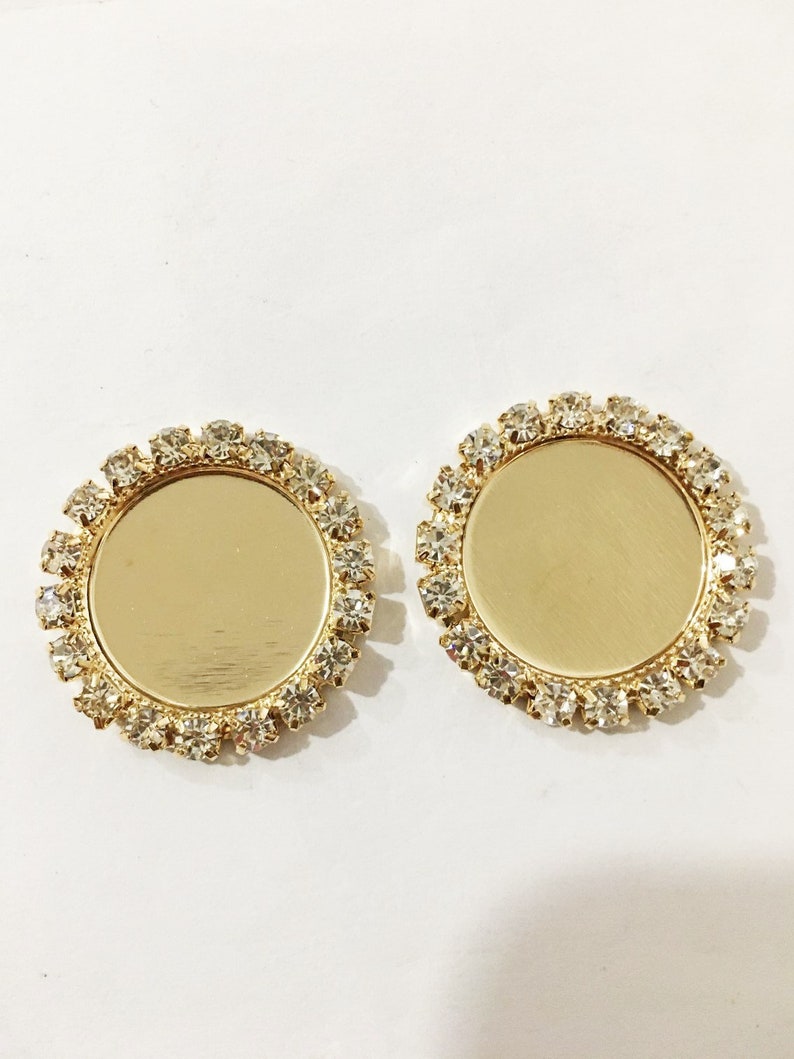May include: Two gold-toned round metal settings with clear rhinestone borders. The settings are blank and can be used for crafting.