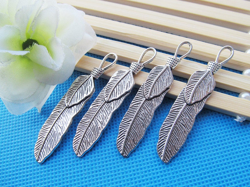May include: Five silver feather charms with a loop at the top for attaching to jewelry or other crafts. The feathers are detailed with a textured surface and have a slightly oxidized finish.