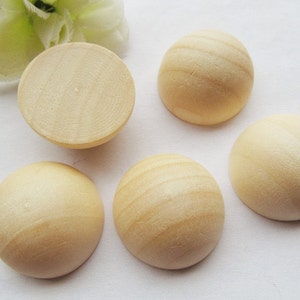 May include: A collection of five unfinished, light-colored wooden half-spheres. One sphere is cut in half, revealing the interior wood grain. The other four are whole, showing the natural wood texture. The spheres are arranged on a white surface.