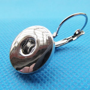 Op de afbeelding: Zilverkleurige oorbel met een ronde knoopvormige hanger en een sluiting met een hendel.