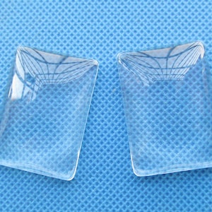 May include: Two clear glass magnifying lenses, rectangular in shape, with rounded corners, on a blue textured surface.