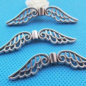 Puede incluir: Tres abalorios de alas de ángel de metal plateado con un diseño de espiral decorativo. Las alas están abiertas y tienen un patrón delicado e intrincado.