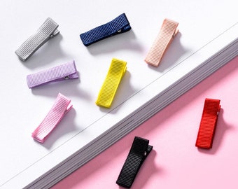 May include: Assortment of colorful hair clips with a satin ribbon finish. The clips are arranged on a white and pink surface.