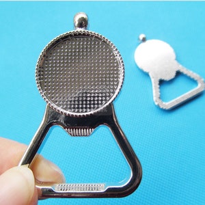 May include: Silver bottle opener with a round, textured top. The opener has a triangular shape with a small loop at the top.