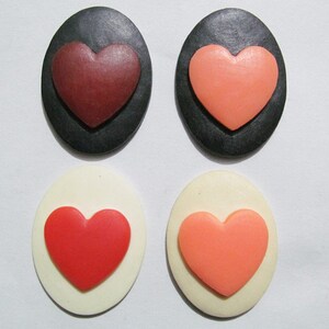 May include: Four oval-shaped resin cabochons with raised heart designs. Two cabochons are black with red and pink hearts. Two cabochons are white with red and pink hearts.