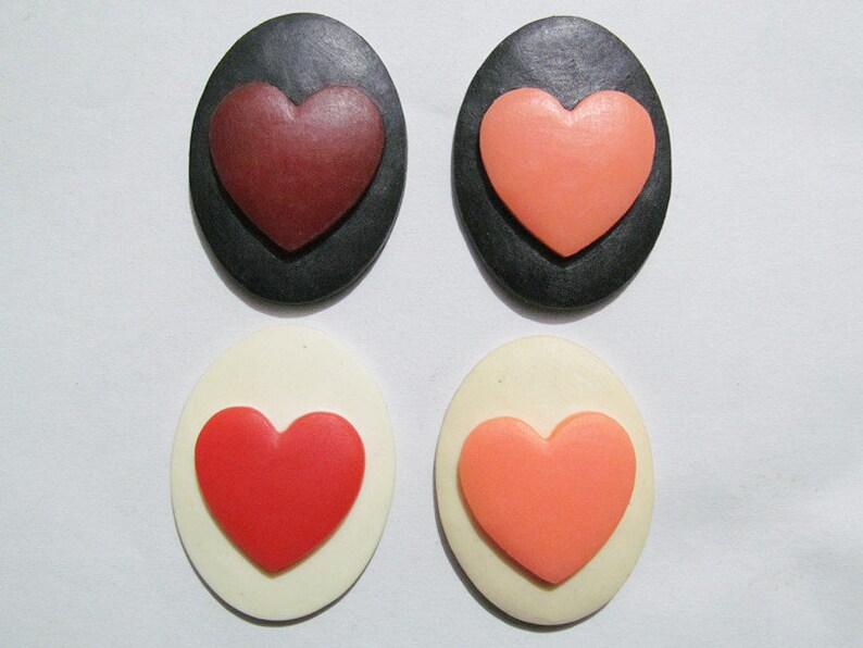 May include: Four oval-shaped resin cabochons with raised heart designs. Two cabochons are black with red and pink hearts. Two cabochons are white with red and pink hearts.