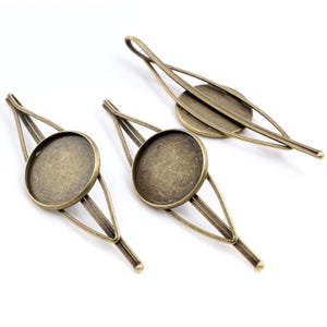 May include: Three antique bronze-coloured hair slides. Each slide has a round, flat setting for embellishments, set within a decorative frame. The slides are arranged on a white surface, showcasing their intricate design.