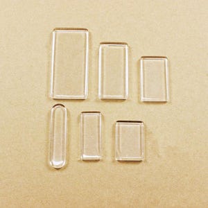 May include: A collection of six clear, smooth, rectangular acrylic or glass cabochons of varying sizes. Five pieces feature sharp, squared corners, while one piece is elongated with rounded ends. Each piece is transparent and flat.