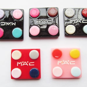 May include: A collection of small, square cosmetic compacts. The compacts are black, red, and pink, each with four circular, colorful accents. The "M·A·C" logo is visible on each compact. The accents are in various shades of pink, blue, yellow, and white.