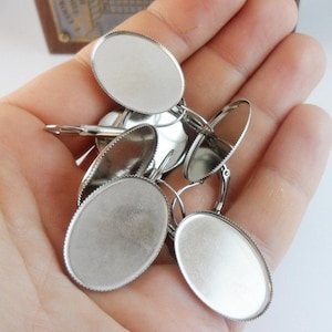 May include: A close-up of a set of silver oval-shaped earring blanks with a scalloped edge. The blanks are in a hand, ready to be used for crafting.