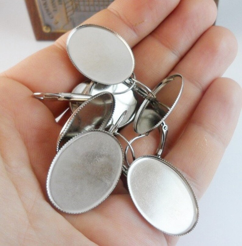 May include: A close-up of a set of silver oval-shaped earring blanks with a scalloped edge. The blanks are in a hand, ready to be used for crafting.