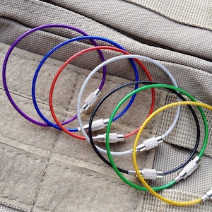 Puede incluir: Un juego de siete llaveros coloridos hechos de cable metálico trenzado. Los llaveros son de varios colores, incluyendo morado, rojo, azul, plateado, verde y amarillo. Los llaveros están dispuestos en círculo sobre un fondo de tela texturizada.