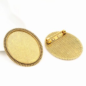 May include: Two gold-coloured oval brooch blanks. One shows the front with a blank surface and a decorative border. The other shows the back with a pin clasp. The brooches are on a white background.