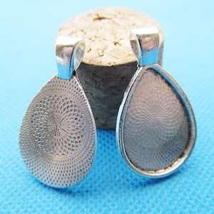 Op de afbeelding: Twee zilverkleurige hanger-bakjes in een traanvorm met een decoratief patroon op de achterkant. De bakjes zijn ontworpen om een platte cabochon te bevatten.