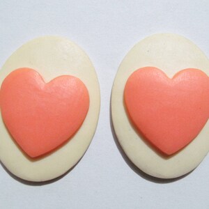 May include: A pair of white oval-shaped earrings with a pink heart design.