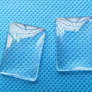 May include: Two clear glass rectangular cabochons, each with a slightly different pattern of light refraction, on a blue textured background.