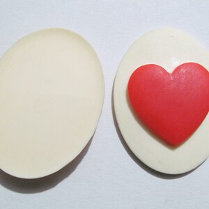 May include: A white oval-shaped trinket box with a red heart-shaped design on the lid. The box is open, revealing an empty interior.