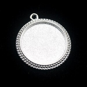 May include: Silver pendant with a round, beveled, and textured edge. The pendant has a smooth, circular surface in the center.
