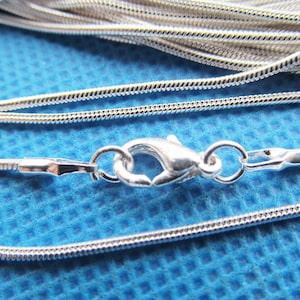 Puede incluir: Un collar de cadena de serpiente de plata con cierre de mosquetón. La cadena está hecha de pequeños eslabones redondos que están estrechamente entrelazados.