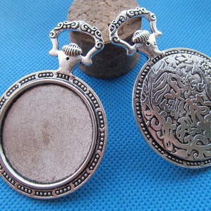 Pode incluir: Dois pendentes em branco de metal prateado com um bezel redondo e uma fivela decorativa. Um pendente tem uma superfície lisa, enquanto o outro tem um design floral intrincado.