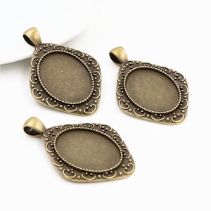 May include: Three antique bronze-colored pendant trays with an oval center and decorative filigree edges. Each pendant has a small loop at the top for attaching to a chain or cord. The pendants are displayed on a white surface.
