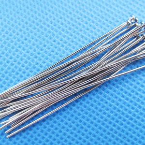 May include: A close-up of a pile of silver-colored straight pins with rounded heads. The pins are arranged in a fan shape on a blue textured surface.