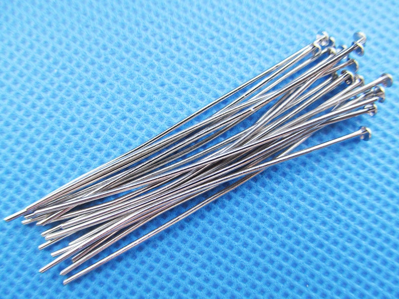 May include: A close-up of a pile of silver-colored straight pins with rounded heads. The pins are arranged in a fan shape on a blue textured surface.