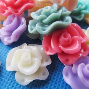 May include: Assortment of colourful plastic flower embellishments in various shades of pink, purple, white, and green. The flowers are all in a similar style, with a ruffled, layered design.