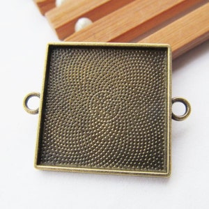 May include: A square, bronze-colored metal frame with two loops on the sides. The frame has a textured surface and is designed to hold a small image or photo.