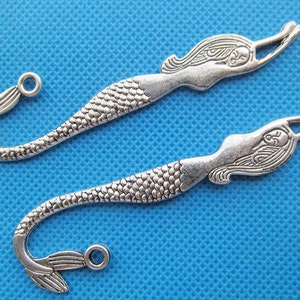 Puede incluir: Dos abalorios de marcapáginas de sirena plateados con un diseño detallado. Cada abalorio presenta una sirena con el pelo suelto y una cola de pez, con un bucle en el extremo de la cola para sujetarlo a un marcapáginas.