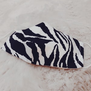 Könnte beinhalten: Eine Gesichtsmaske mit einem schwarz-weißen Zebrastreifenmuster. Die Maske hat weiße elastische Ohrschlaufen und ist aus Stoff gefertigt. Die Maske liegt auf einer weißen, flauschigen Oberfläche.