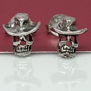May include: A pair of silver skull earrings with cowboy hats. The skulls have detailed features and the hats are slightly tilted.