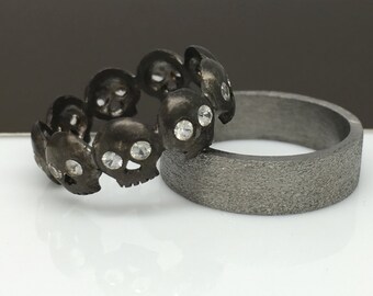 May include: Two black metal rings. One ring is a band with a textured surface. The other ring is a band of skulls with clear gemstones in the eye sockets.