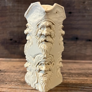 May include: A carved wooden sculpture featuring the faces of bearded men wearing hats. The sculpture is rectangular and has multiple faces carved into its sides. The wood has a natural, light color and a textured surface.