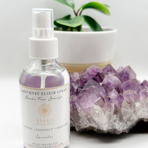 May include: A clear glass bottle with a white spray nozzle and a white label. The label says "Amethyst Elixir Spray Smoke-Free Smudge" and "Shakti Healing Space". The label also says "Cleanse - Connect - Restore" and "Lavender". The bottle is sitting on a cluster of amethyst crystals.