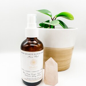 May include: A brown glass bottle with a white spray top and a rose quartz crystal. The bottle is labeled "Rose Quartz Elixir Spray Aura Mist" and "Shakti Healing Space". The label also lists the ingredients: Blue Tansy, Orange Tangerine, Patchouli, Ylang Ylang. The text "LOVE - LIGHT - HEALING - PEACE" is printed on the label.