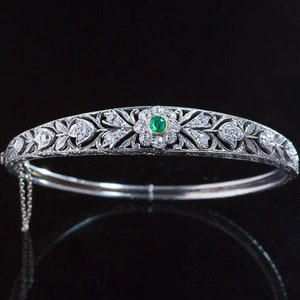Op de afbeelding: Een delicate zilveren armband met een bloemmotief en een enkele smaragdgroene edelsteen in het midden.