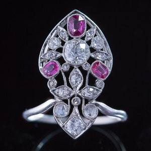 Puede incluir: Un anillo de plata con un gran diamante redondo en el centro, rodeado de diamantes más pequeños y rubíes rosas. El anillo está engastado en un diseño floral.