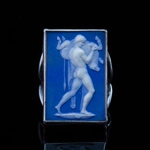 May include: A rectangular ring with a blue cameo depicting a classical figure carrying an animal. The ring has a silver-coloured metal band. The cameo design is white and the background is a deep blue.