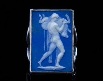 Può includere: Anello rettangolare con cammeo blu raffigurante una figura classica che trasporta un animale. L'anello ha una fascia in metallo color argento. Il design del cammeo è bianco e lo sfondo è blu intenso.
