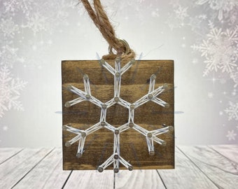 Peut inclure: Une décoration de flocon de neige en bois avec une ficelle blanche enroulée autour de clous. L'ornement a un cintre en ficelle brune.