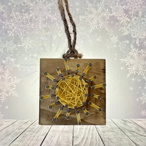 May include: A yellow string art sun ornament on a wooden square with a twine hanger. The sun is made with yellow string wrapped around nails hammered into the wood.