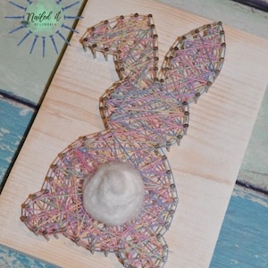 Puede incluir: Una pieza de arte de hilo de un conejo rosa y azul con una cola de pompón blanco. El conejo está hecho con hilo envuelto alrededor de clavos martillados en una tabla de madera. La tabla es de color marrón claro y tiene el texto "Nailed it by Y'know What" en la esquina superior izquierda.