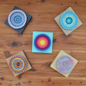 May include: Five colorful string art pieces on wooden squares. Each piece features a circular design with different colors and patterns. The designs include a rainbow, blue, purple, and pink.
