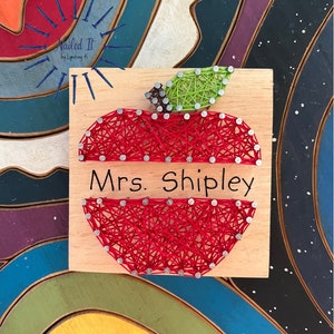 May include: A red apple with a green leaf is made with string wrapped around nails on a wooden board. The apple has the text "Mrs. Shipley" written on it. The board has a blue and white star pattern in the background.