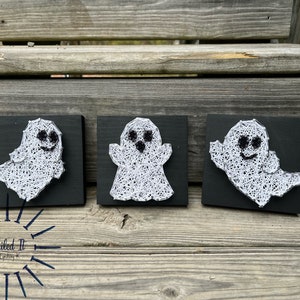 May include: Three small wooden squares with black backgrounds and white string art ghosts with black eyes and mouths. The squares are arranged horizontally on a wooden surface.