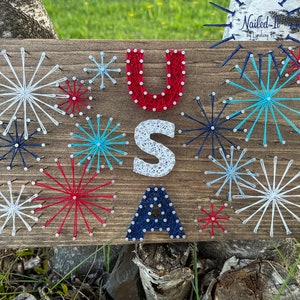 May include: A wooden sign with the letters "USA" spelled out in string art. The letters are surrounded by red, white, and blue string art fireworks.